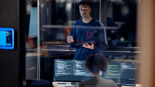 Man Holding Tablet Talking To Woman At Computer 