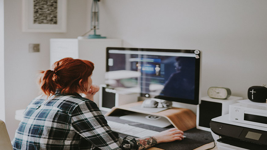 A young person wearing a plaid shirt with their red hair in a ponytail working on a Mac computer monitor.